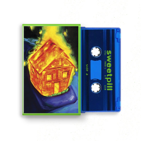 A blue cassette tape from Sweet Pill lies beside its case, which displays vibrant art of a wooden house on fire against a dark backdrop, echoing the limited edition vinyl’s unique style and exclusive tracks.