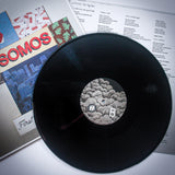 A Somos black vinyl record labeled B sits on its sleeve, with SOMOS First Day Back cover art and a printed lyric sheet partially visible beneath, reminiscent of a black backpack on the first day back to school.