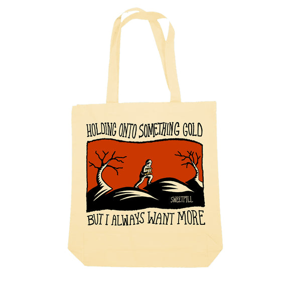 A beige heavy cotton tote by Sweet Pill showcases an exclusive design: a person holding something on a hill with bare trees, an orange background, and the text “Holding onto something gold but I always want more.”.